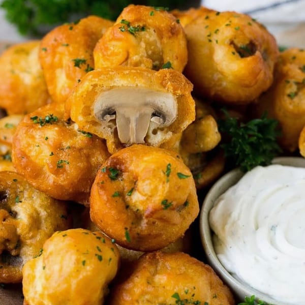 Golden-brown fried mushrooms, one cut in half, with a side of creamy white dipping sauce and parsley.